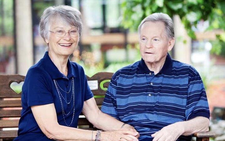 A senior woman and a senior man sitting together on a brown wooden bench outdoors at a park